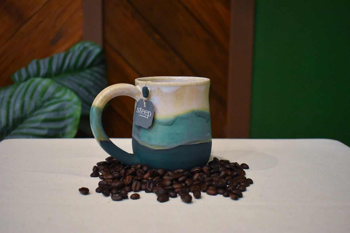 Hand-thrown mug in Lakeshore, warm cream with a gold line over soft aqua and deep matte teal