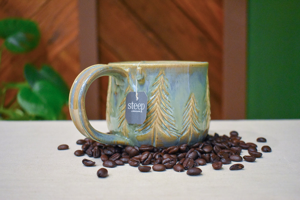 Hand-thrown mug carved with a stand of pine trees in Ivy Pines, honey-gold branches on soft sage and blue-grey