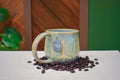 Hand-thrown mug carved with a stand of pine trees in Ivy Pines, honey-gold branches on soft sage and blue-grey