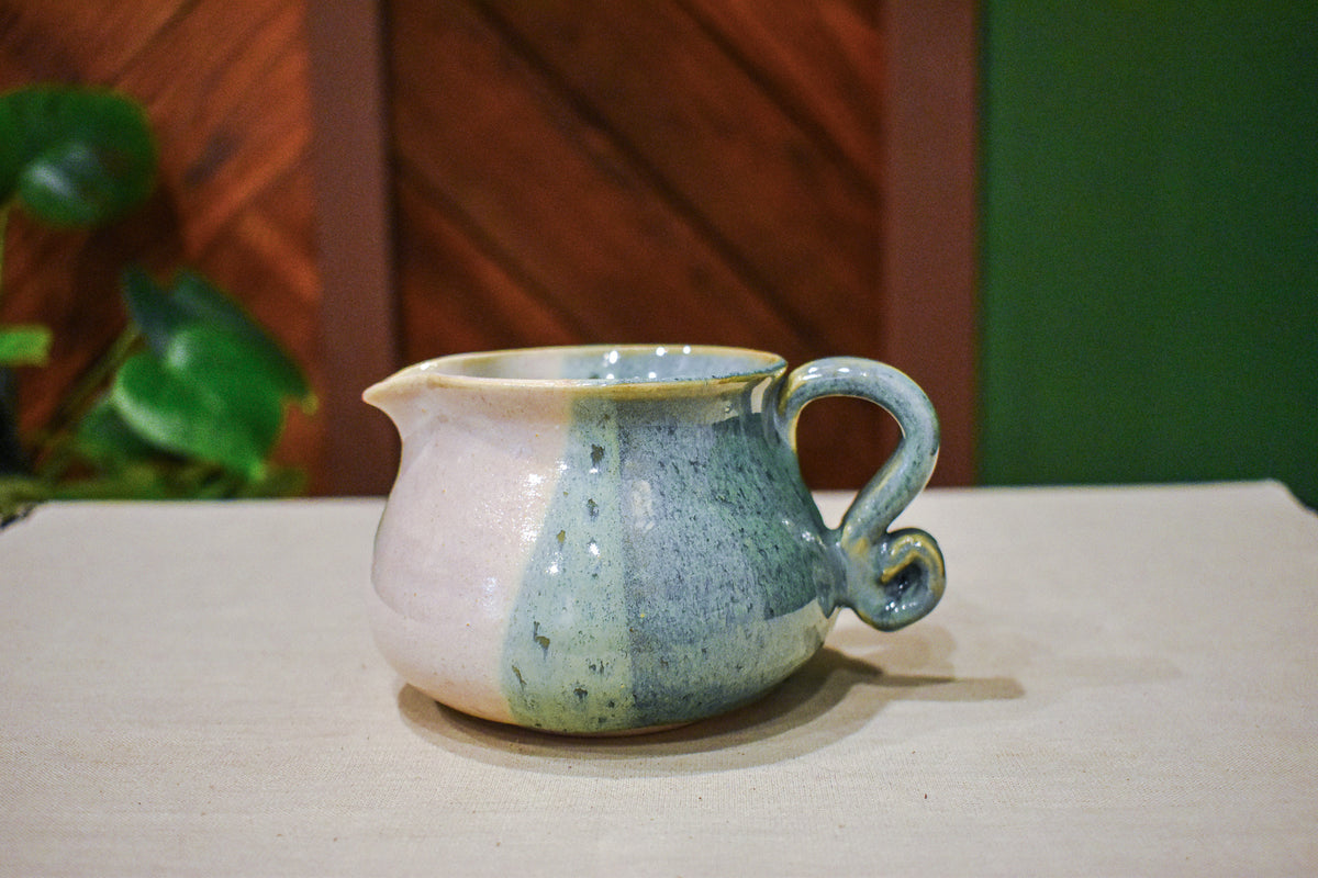 Hand-thrown creamer with a pour spout and curled handle, sage green on one side and warm cream on the other