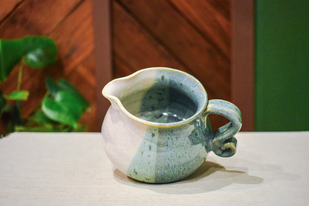 Hand-thrown creamer with a pour spout and curled handle, sage green on one side and warm cream on the other