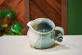 Hand-thrown creamer with a pour spout and curled handle, sage green on one side and warm cream on the other