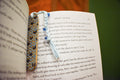 Hand-formed stoneware bookmark with a sky-blue knit-stitch texture, navy stitches and a white braided cord with blue beads