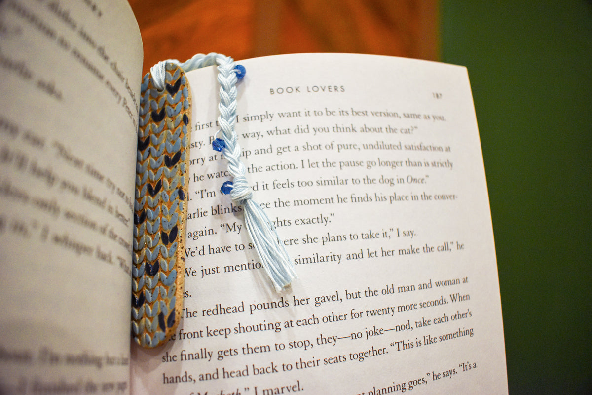 Hand-formed stoneware bookmark with a sky-blue knit-stitch texture, navy stitches and a white braided cord with blue beads