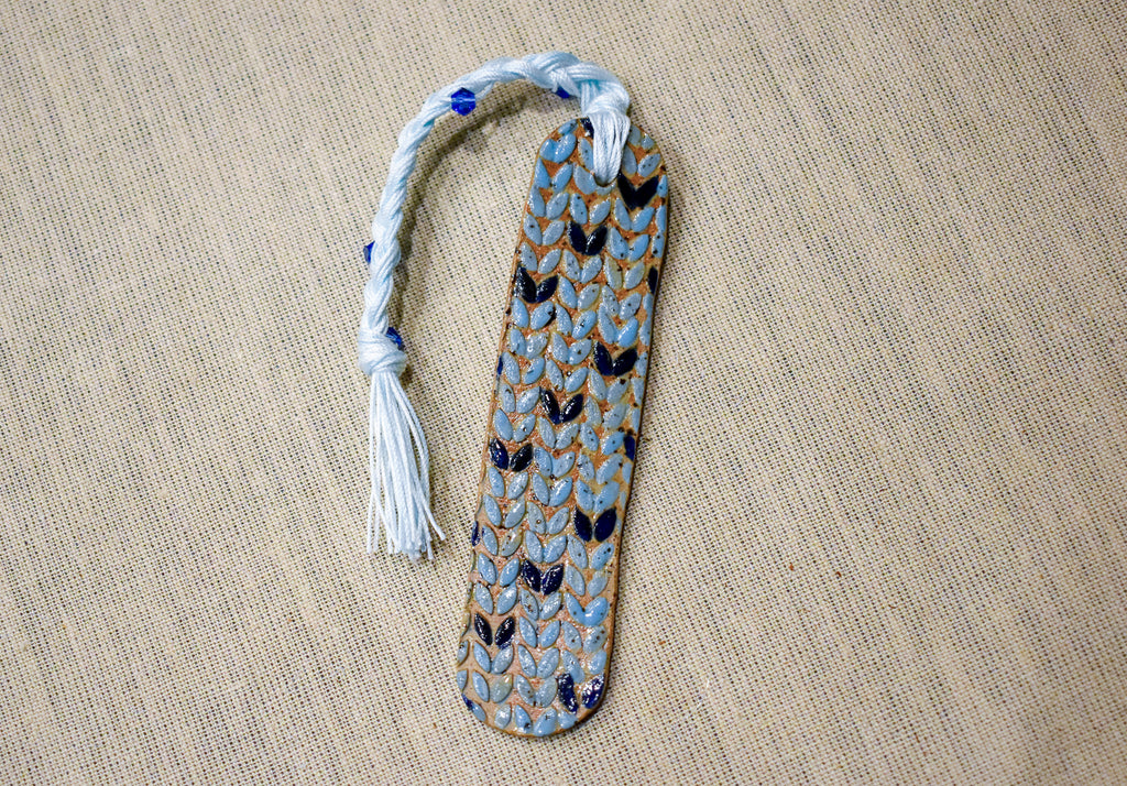 Hand-formed stoneware bookmark with a sky-blue knit-stitch texture, navy stitches and a white braided cord with blue beads